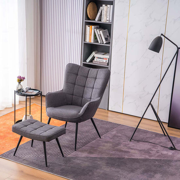 A charcoal linen accent chair with a matching footstool, both with black tapered legs, positioned in a room with a floor lamp, side table, and bookshelf.