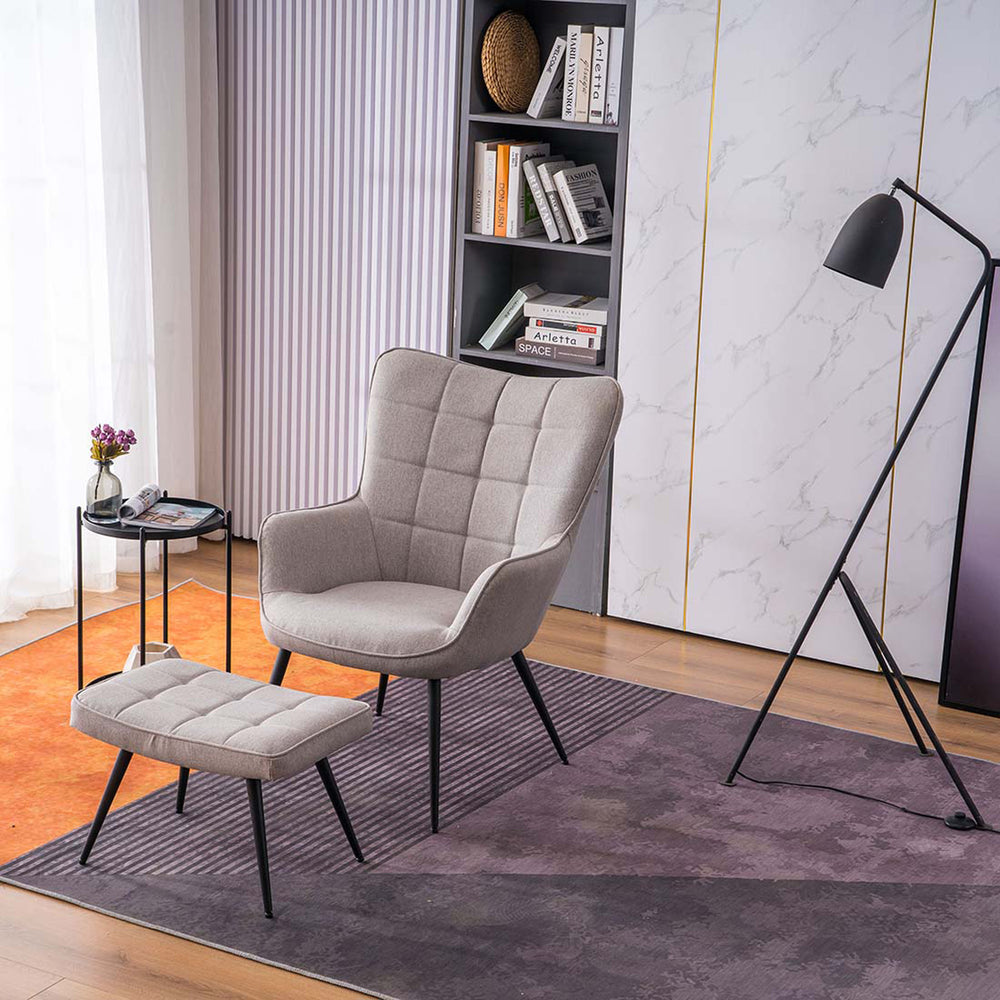 A light grey linen accent chair with a matching footstool, both with black tapered legs, positioned in a room with a floor lamp, side table, and bookshelf.