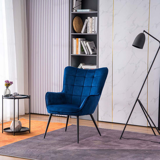 A blue velvet accent chair with black tapered legs, placed in a room with a round side table, a floor lamp, and a bookshelf filled with books and decorative items.