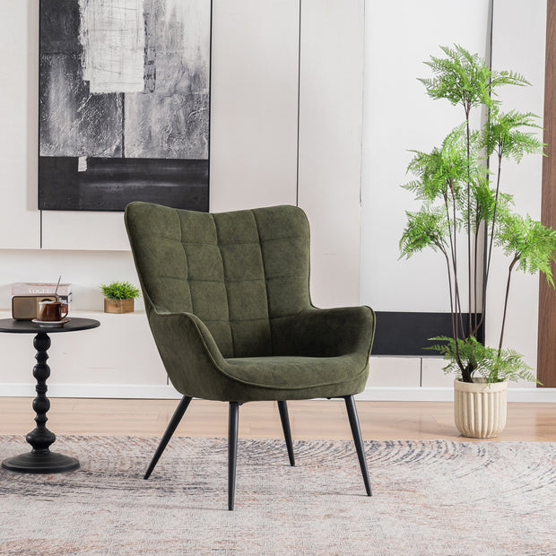 A green velvet accent chair with black tapered legs, placed in a room with a round side table, a potted plant, and a large abstract painting on the wall.
