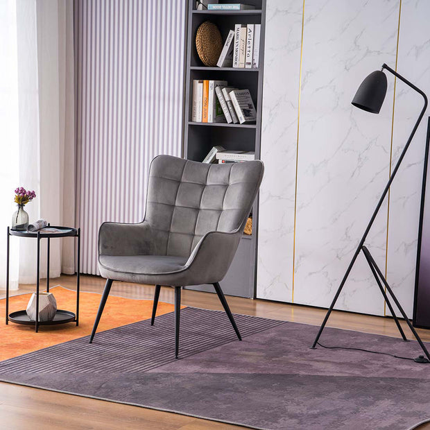 A grey velvet accent chair with black tapered legs, placed in a room with a round side table, a floor lamp, and a bookshelf filled with books and decorative items.