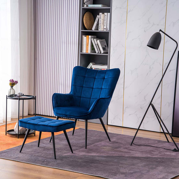 A blue velvet accent chair with a matching footstool, both with black tapered legs, positioned in a room with a floor lamp, side table, and bookshelf.