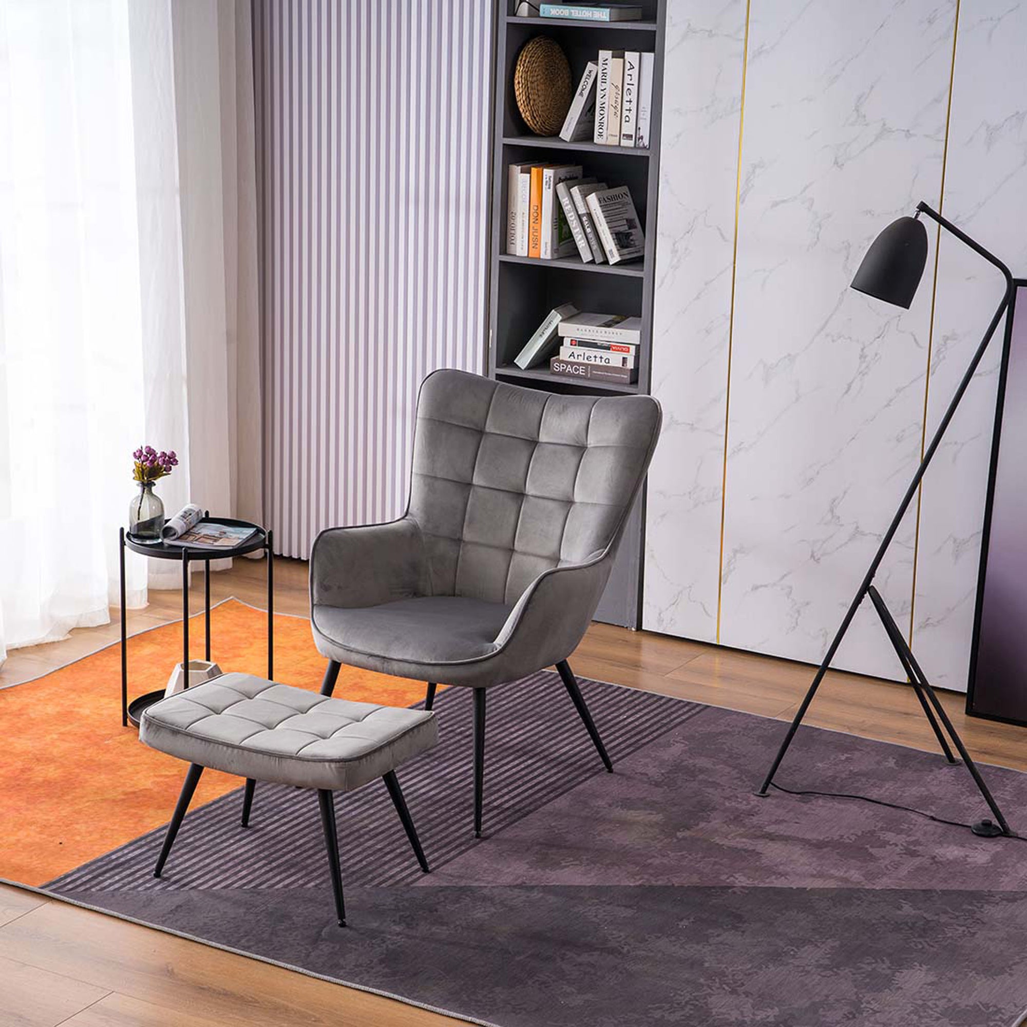 A grey velvet accent chair with a matching footstool, both with black tapered legs, positioned in a room with a floor lamp, side table, and bookshelf.
