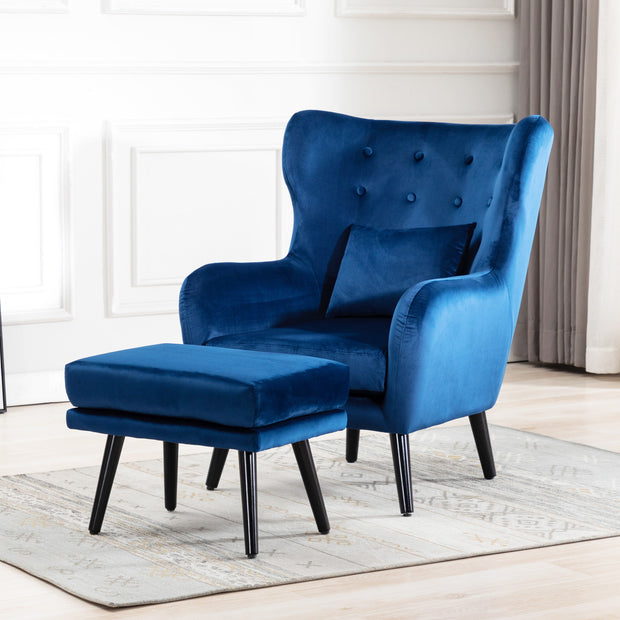 Blue velvet accent wingback chair with tufted buttons, paired with a matching footstool, set on a grey patterned rug against a white panelled wall.