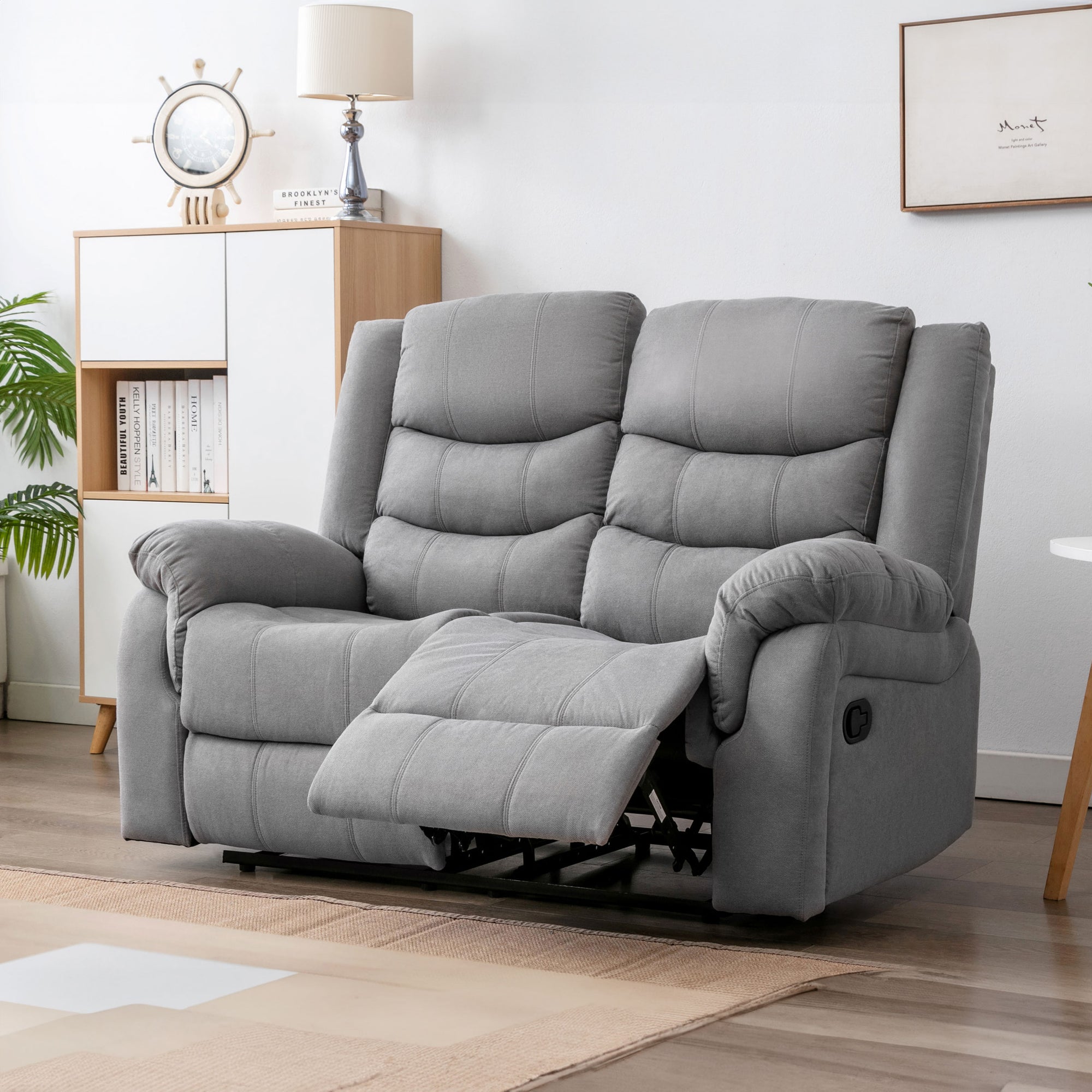 Grey fabric two-seater manual reclining sofa with one seat extended, placed in a cosy living room with light wooden furniture, a nautical clock, and framed artwork.