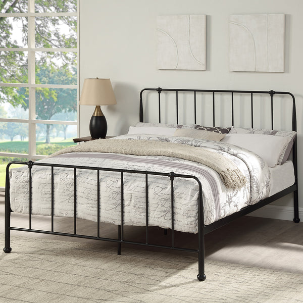 Black metal bed frame with vertical bars on the headboard and footboard. The bed has beige bedding with script patterns and several pillows. A lamp on a bedside table and two abstract wall hangings are in the background.