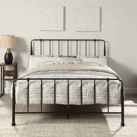 Black metal bed frame with vertical bars on the headboard and footboard. The bed has beige bedding with script patterns and several pillows. A lamp on a bedside table and two abstract wall hangings are in the background.