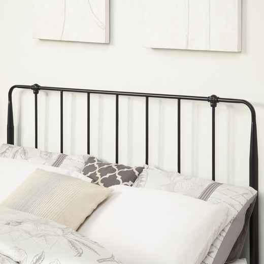 Close-up of a black metal headboard with vertical bars. The bed is made with patterned bedding and pillows. Two abstract wall hangings are visible in the background against a light-colored wall.