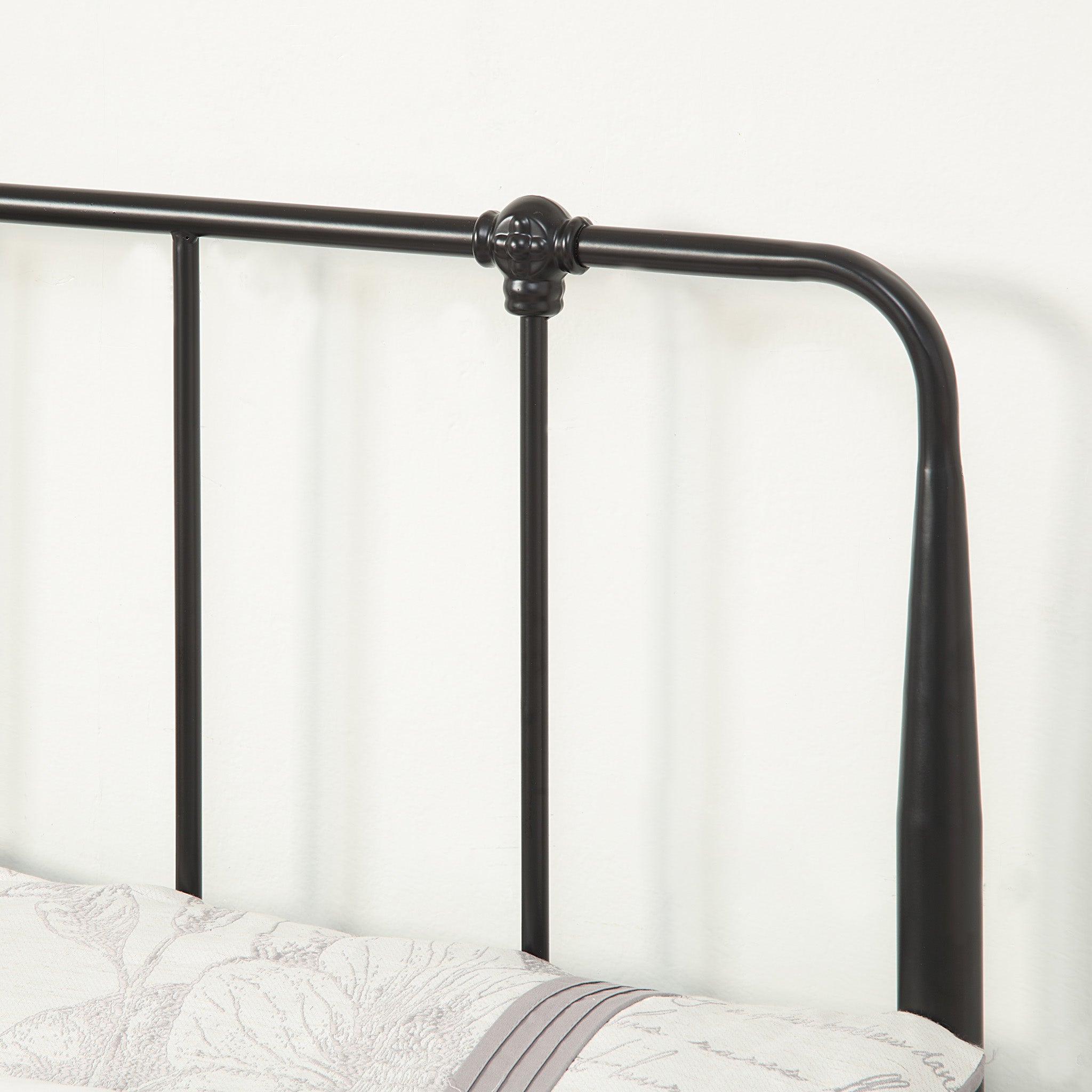 Close-up of a black metal headboard with vertical bars. The bed is made with patterned bedding, and the background is a plain light-colored wall.