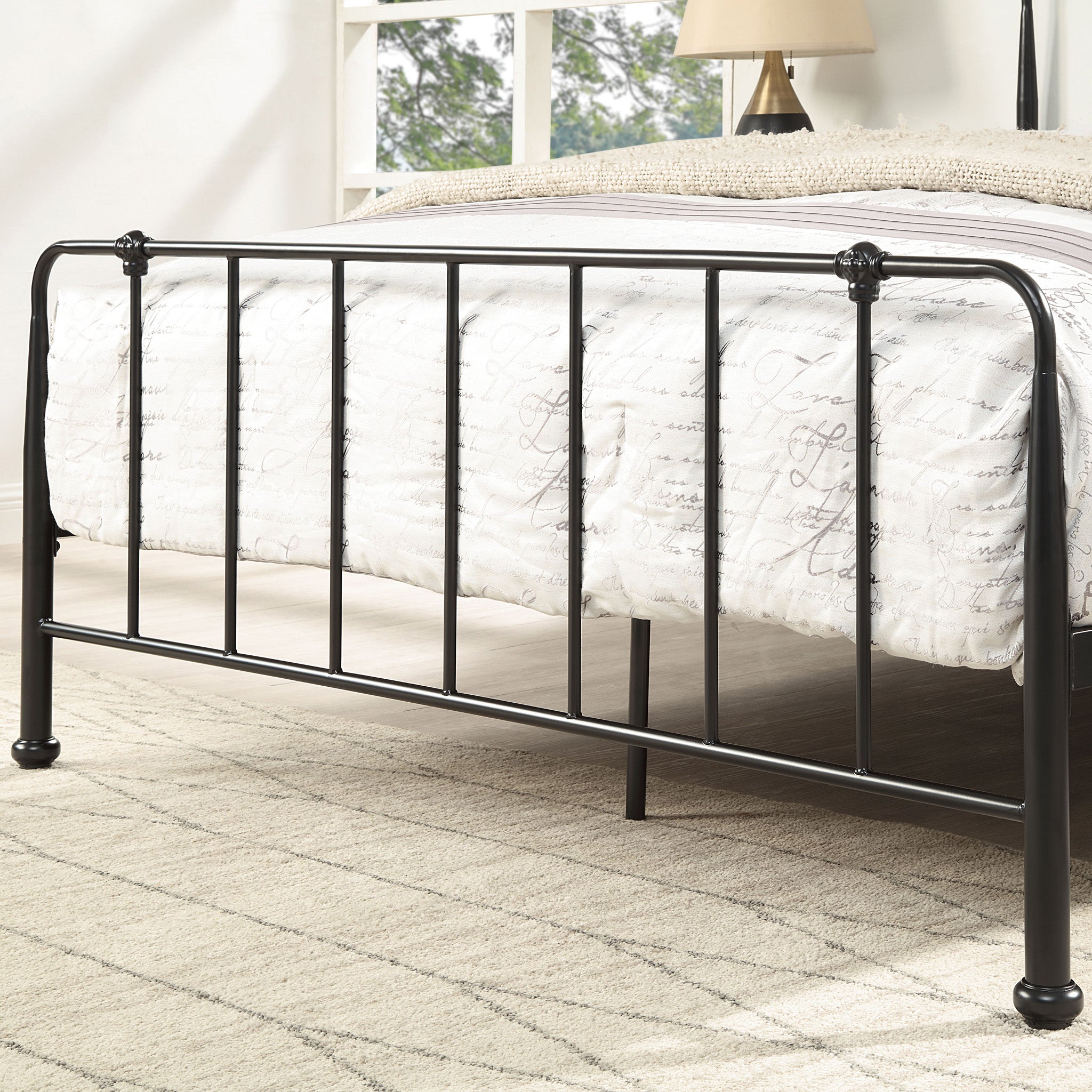 Close-up of a black metal footboard with vertical bars. The bed is covered with white bedding featuring script patterns. A window, lamp, and light-colored carpet are in the background.