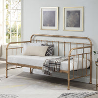 A brass-coloured metal day bed is adorned with white bedding, decorative pillows, and a plaid throw blanket. Two paintings hang above the bed.