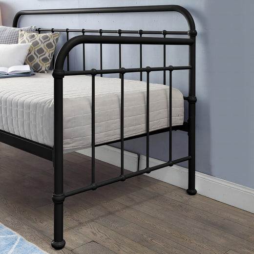 A close-up view of the side of a black metal bed frame with intricate detailing, featuring a white mattress and decorative cushions, set against a grey wall and wooden floor.