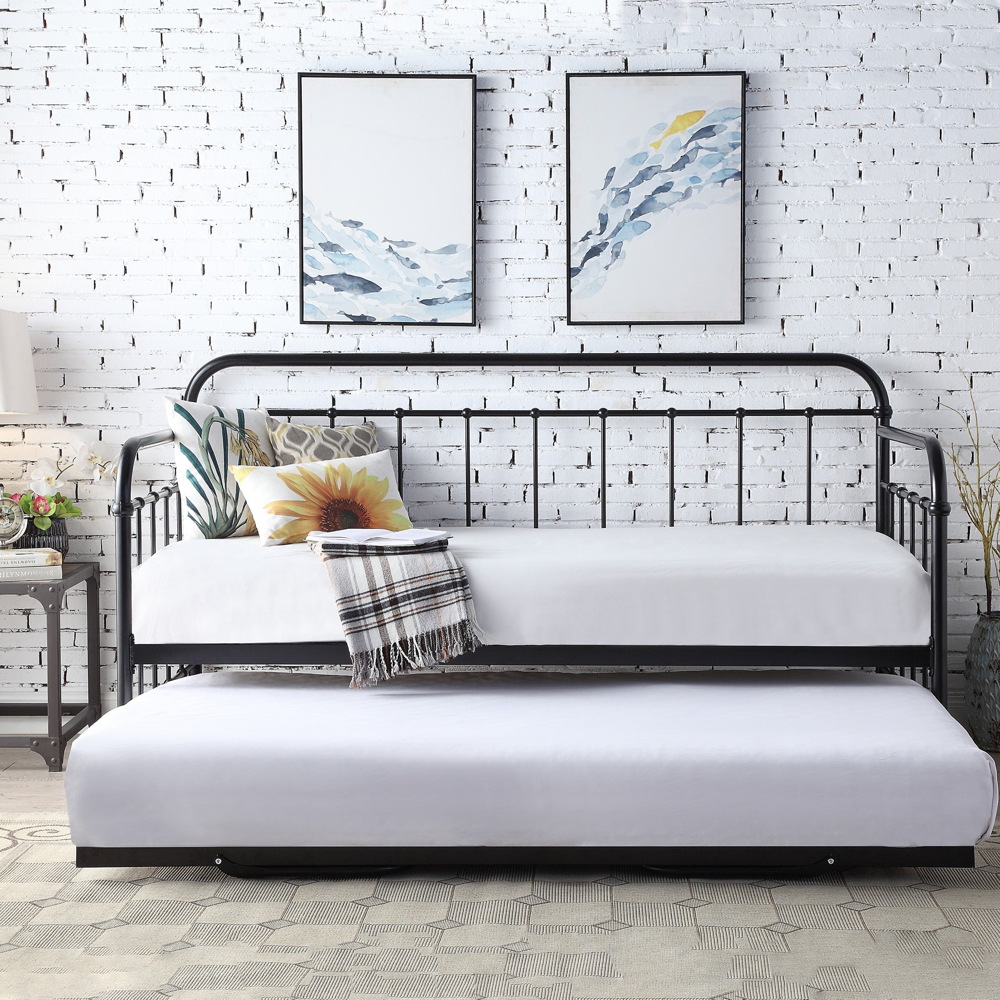 Black metal daybed with a pull-out trundle, white linens, and decorative pillows, set against a white brick wall.