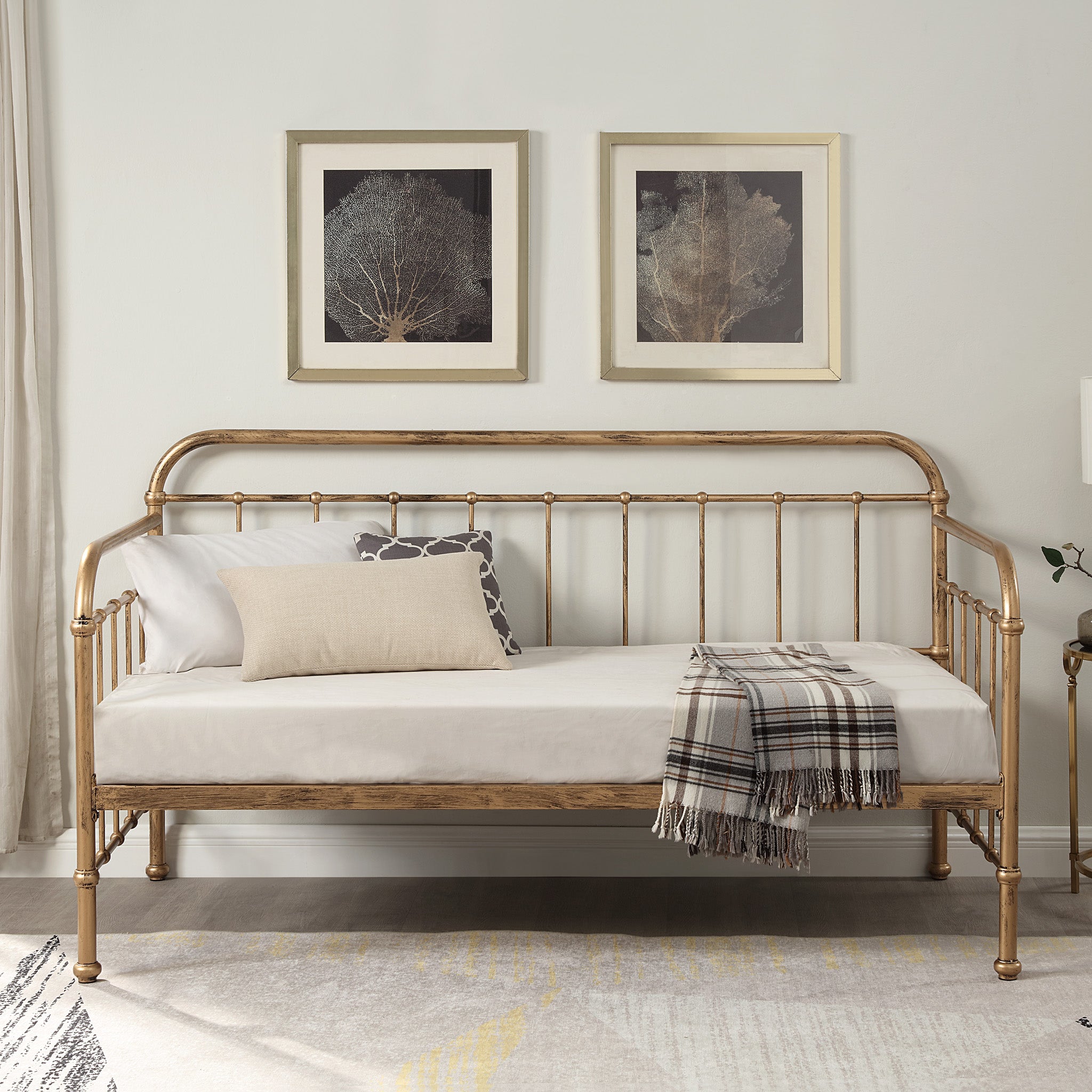 A brass-coloured metal day bed is adorned with white bedding, decorative pillows, and a plaid throw blanket. Two paintings hang above the bed.