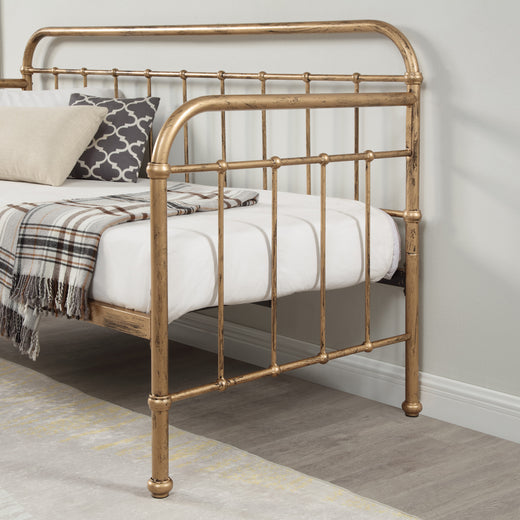 A close-up view of a brass-coloured metal bed frame with intricate detailing, featuring a white mattress and a plaid throw blanket, set against a white wall and wooden floor.