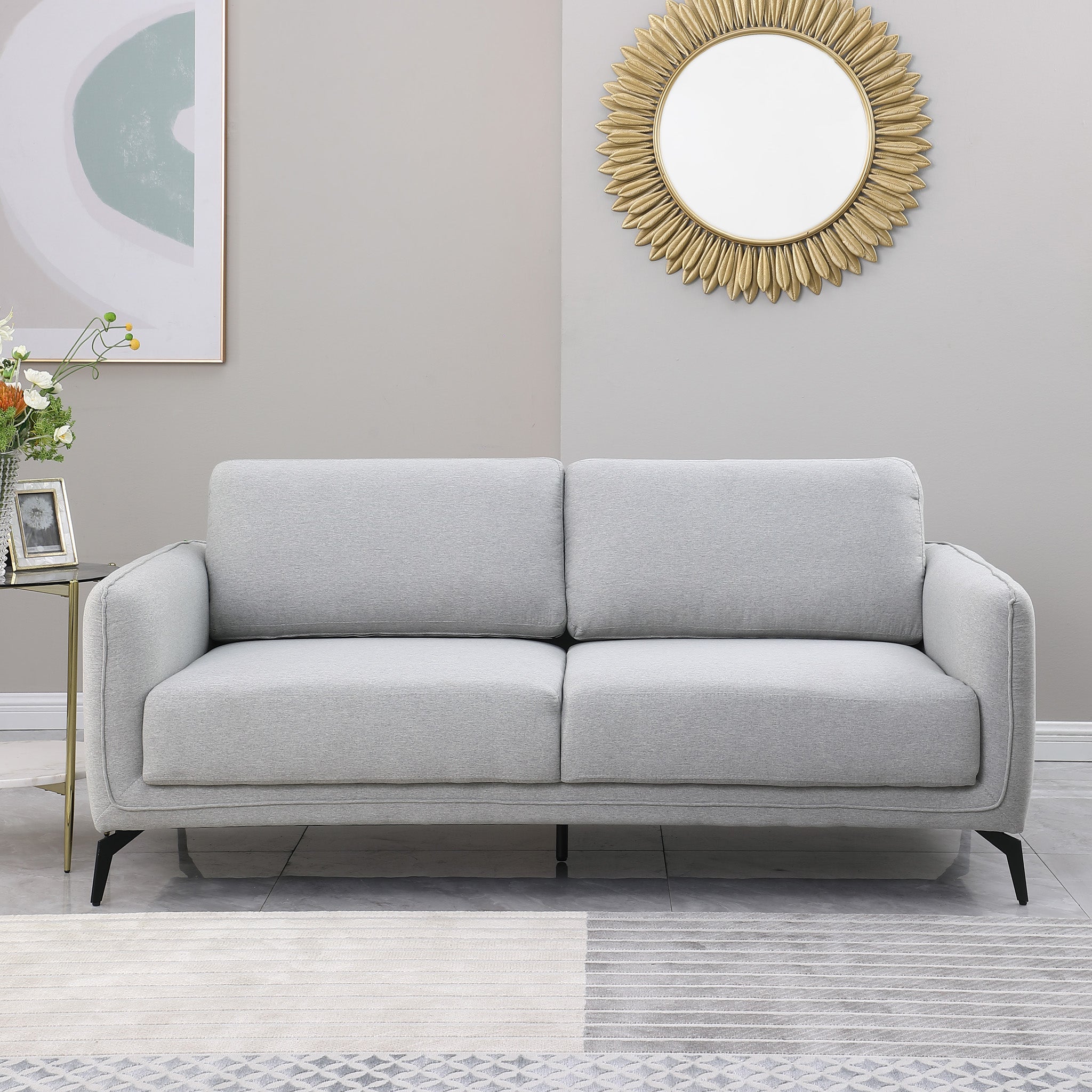 Light grey upholstered sofa with black metal legs in a grey living room, featuring a round sunburst mirror and a side table with flowers.