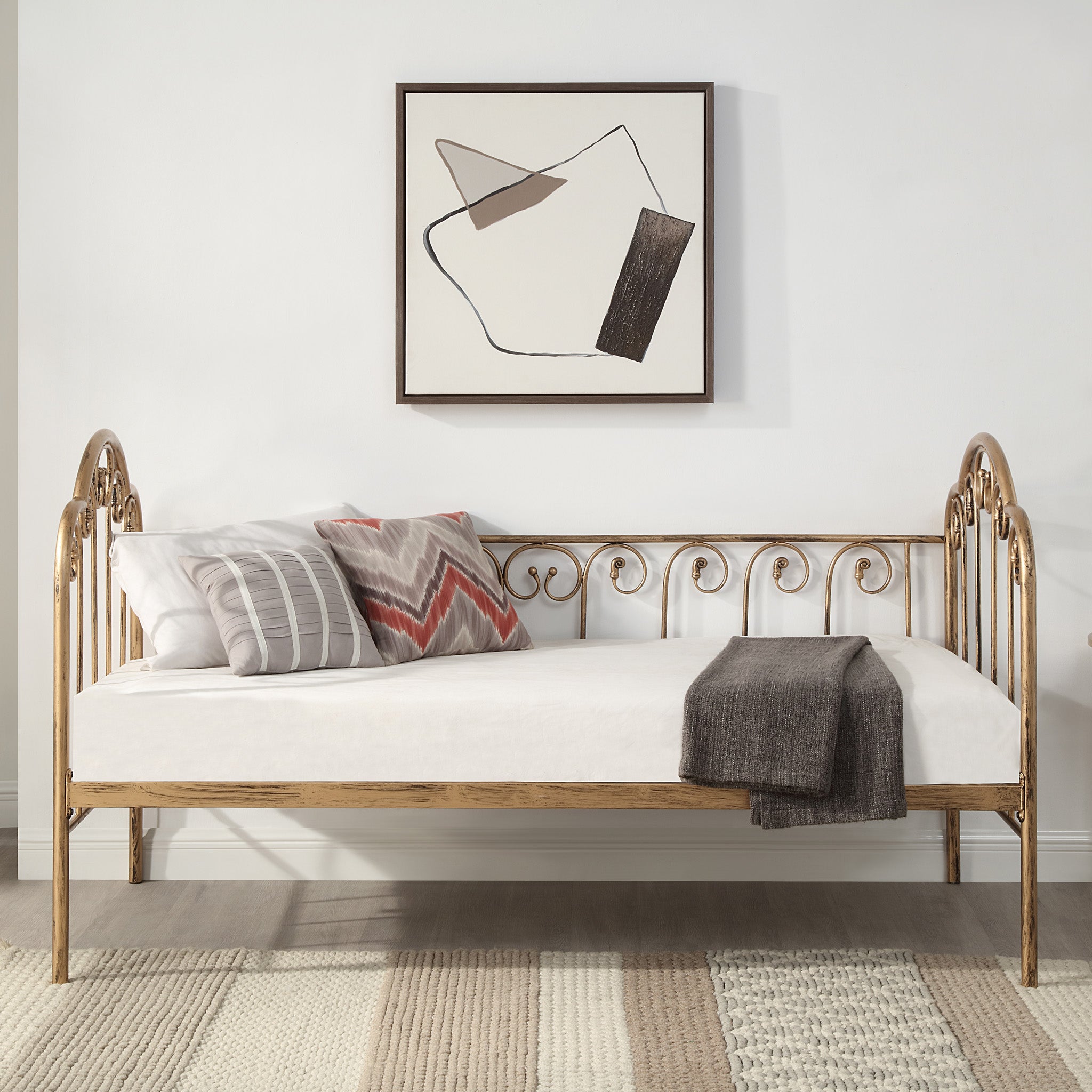 A brass metal day bed is adorned with white bedding, decorative pillows, and a grey throw blanket. A geometric painting hangs above the bed.