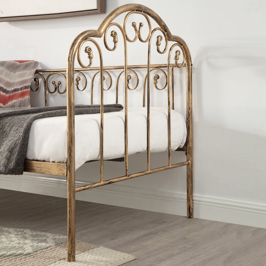 A close-up view of a brass-coloured metal bed frame with intricate detailing, featuring a white mattress and a dark grey throw blanket, set against a white wall and wooden floor.