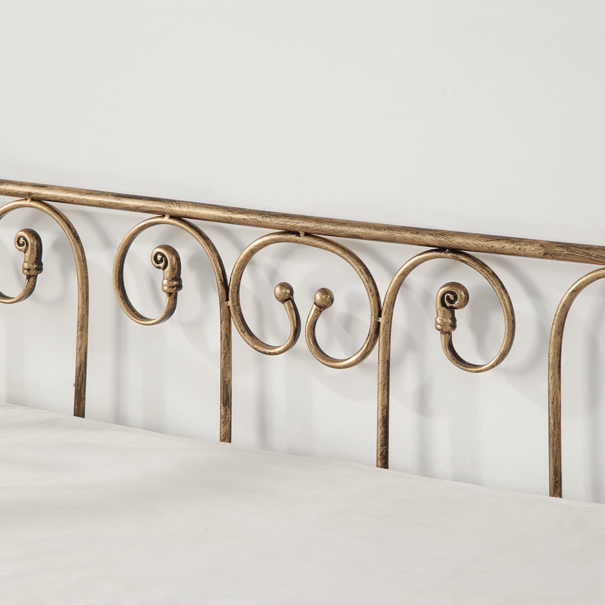 Close-up view of a brass-coloured metal bedframe, featuring scrollwork, against an off-white wall.