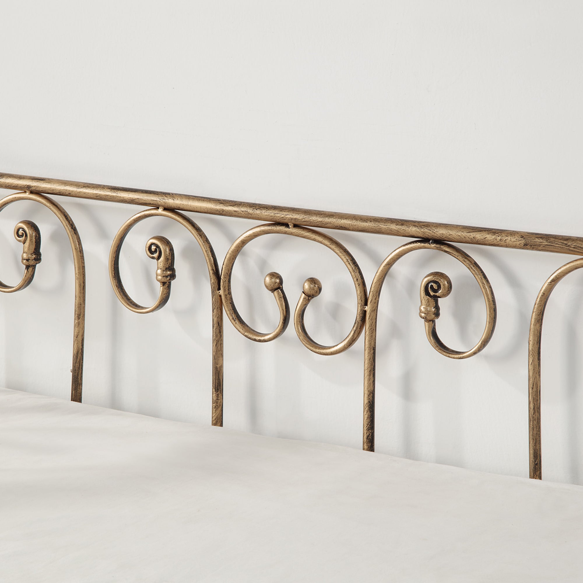 Close-up view of a brass-coloured metal bedframe, featuring scrollwork, against an off-white wall.