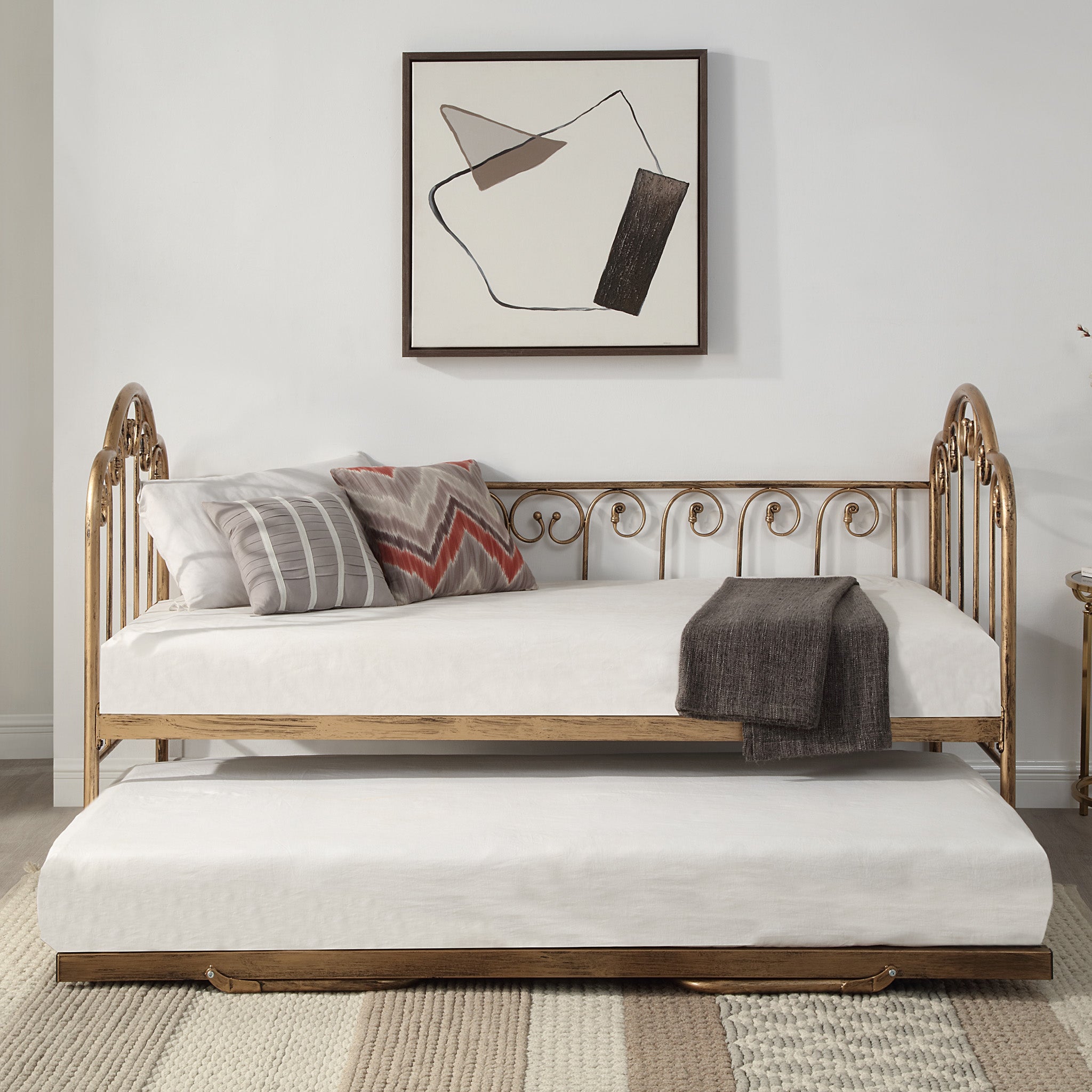  A brass metal day bed with an extended trundle is adorned with white bedding, decorative pillows, and a grey throw blanket. A geometric painting hangs above the bed. 