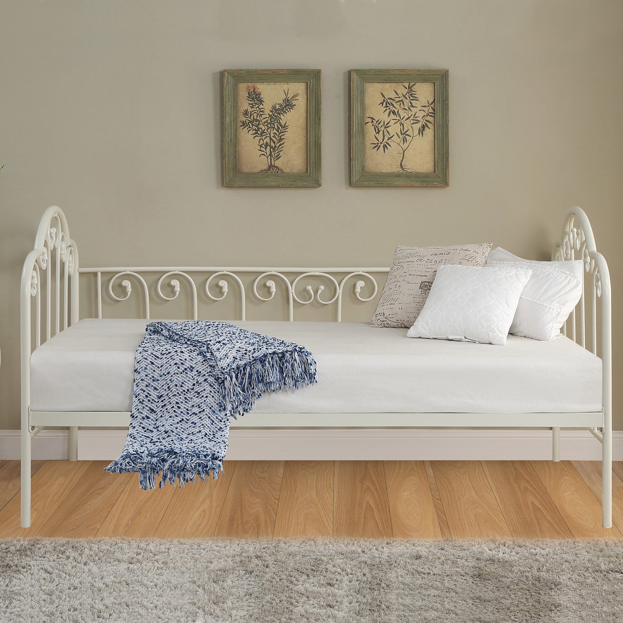 A white metal day bed is adorned with white bedding, decorative pillows, and a blue throw blanket. Two botanical paintings hang above the bed on a beige wall. 