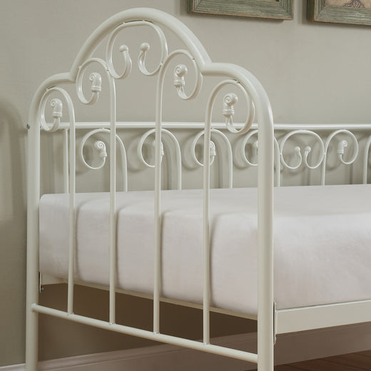 A close-up view of a white metal bed frame with intricate detailing, featuring a white mattress, set against a beige wall.