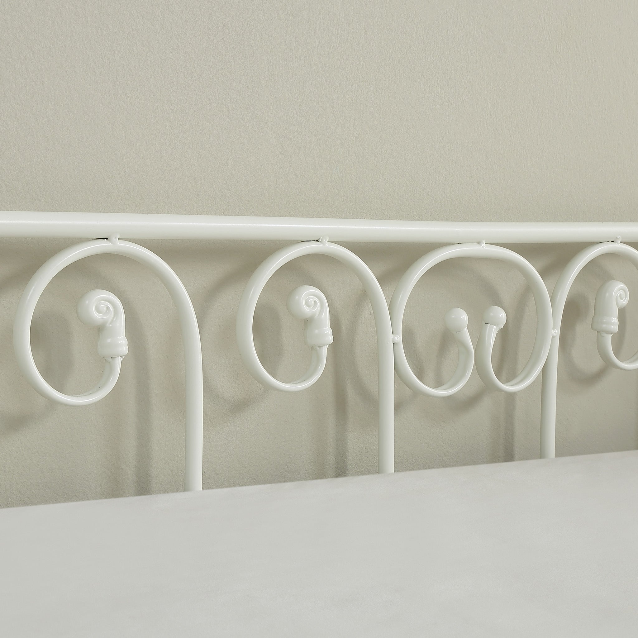 Close-up view of a white metal bedframe, featuring scrollwork, against an off-white wall.