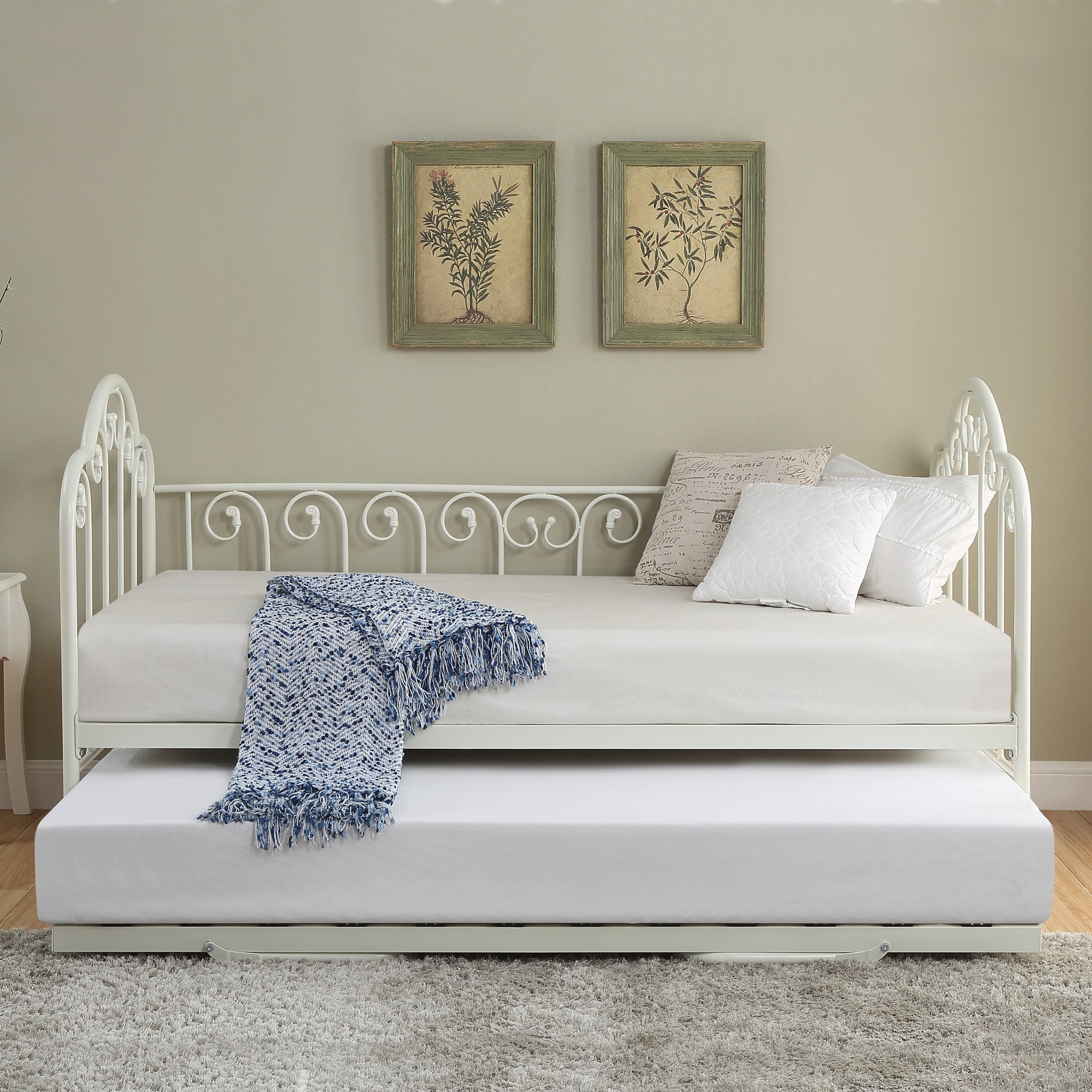  A white metal day bed with an extended trundle is adorned with white bedding, decorative pillows, and a blue throw blanket. Two botanical prints hang above the bed.