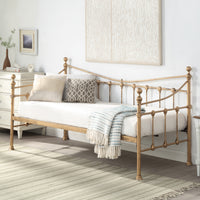 A brass-coloured metal day bed is adorned with white bedding, decorative pillows, and a light blue throw blanket. A painting hangs above the bed on a white wall.