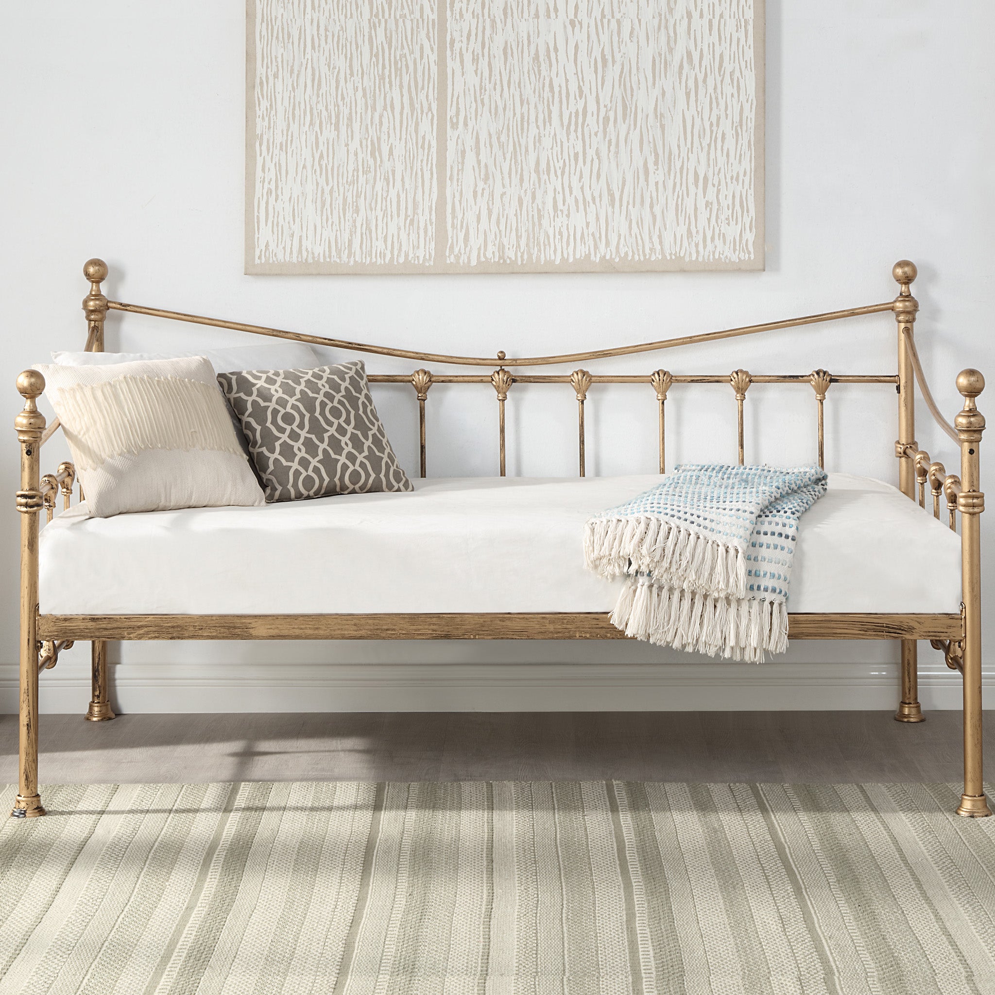 A brass-coloured metal day bed is adorned with white bedding, decorative pillows, and a light blue throw blanket. A painting hangs above the bed on a white wall.