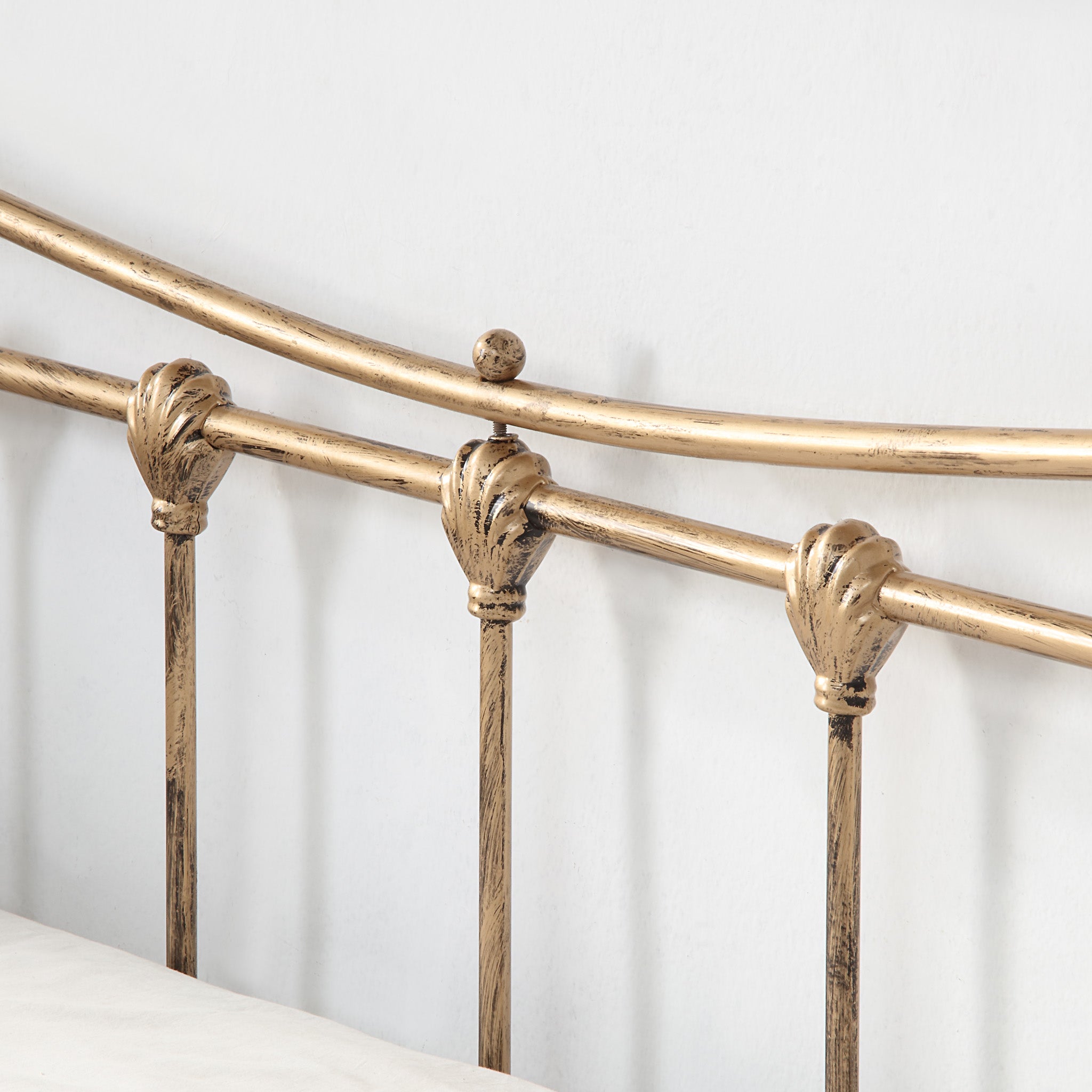 A close-up view of the detailing on a brass-coloured metal bed frame, highlighting the curved design elements against a white wall