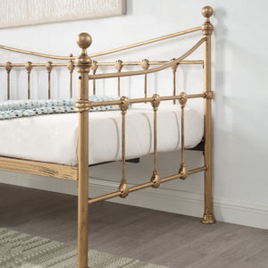 A close-up view of a brass-coloured metal bed frame with intricate detailing, featuring a white mattress and a light blue patterned blanket, set against a white wall and wooden floor.