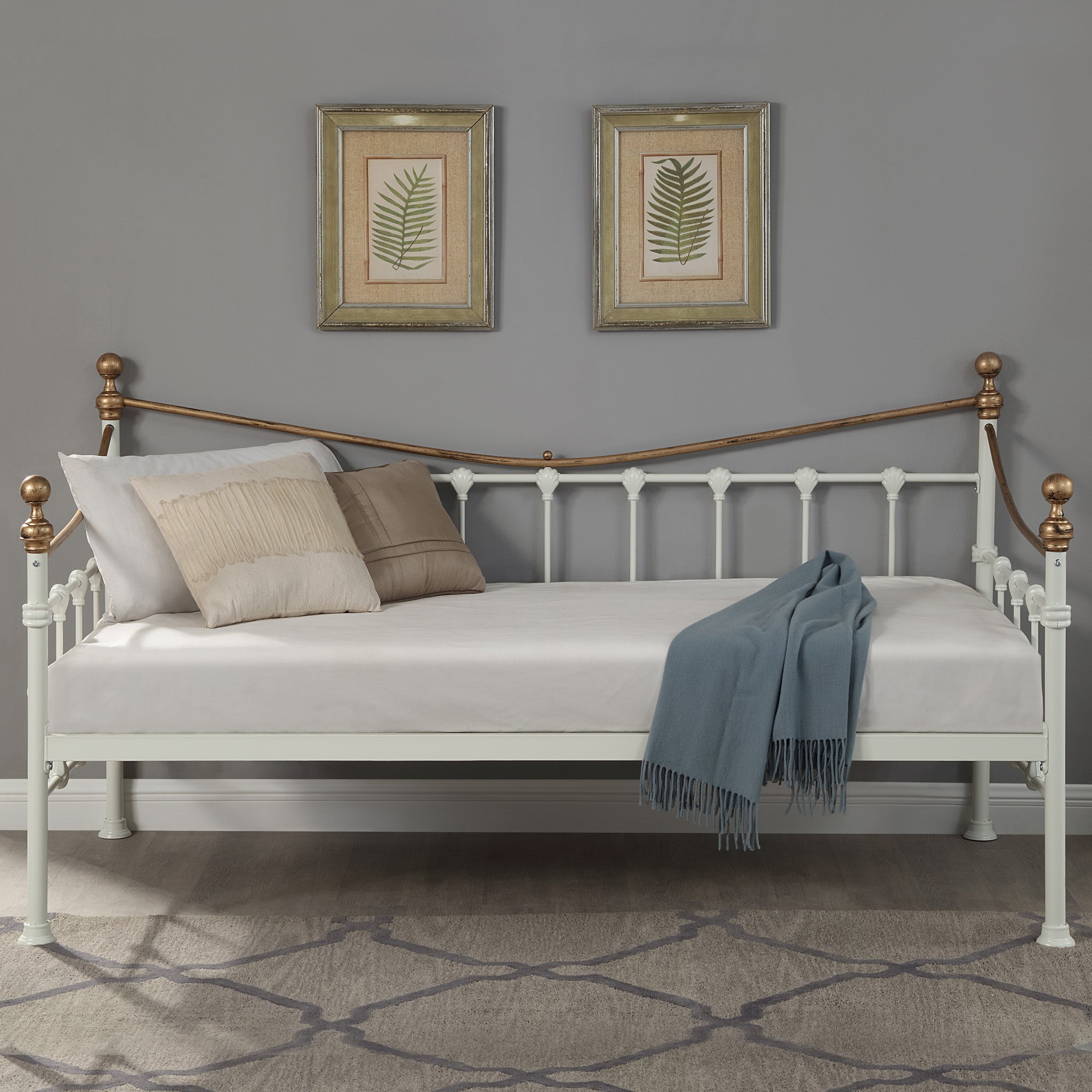 A brass and white metal day bed is adorned with white bedding, decorative pillows, and a light grey throw blanket. Two botanical paintings hang above the bed on a grey wall.