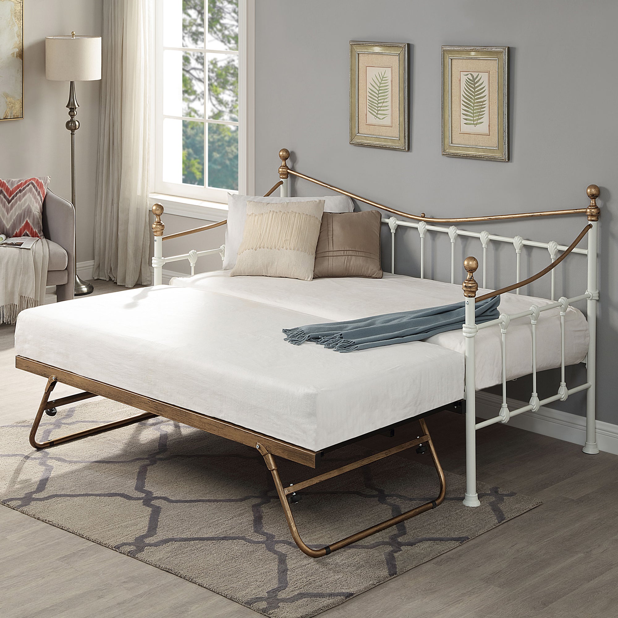 A brass and white metal day bed with an open trundle is adorned with white bedding, decorative pillows, and a light grey throw blanket. Two botanical paintings hang above the bed. 