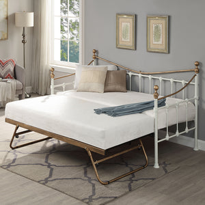 A brass and white metal day bed with an open trundle is adorned with white bedding, decorative pillows, and a light grey throw blanket. Two botanical paintings hang above the bed. 