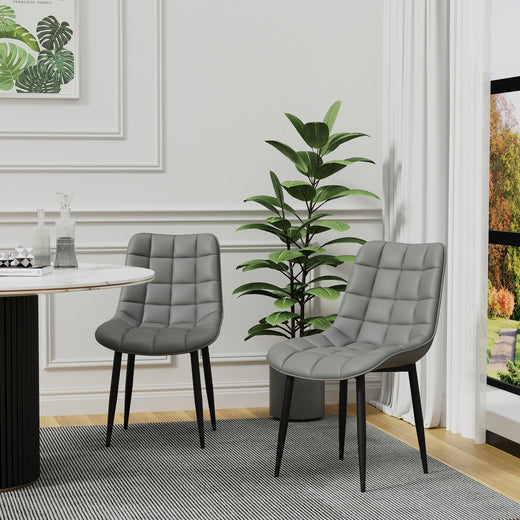 Two grey tufted leather chairs with black legs on a gray patterned rug. A tall green plant, a round dining table, and white walls with decorative molding are in the background.