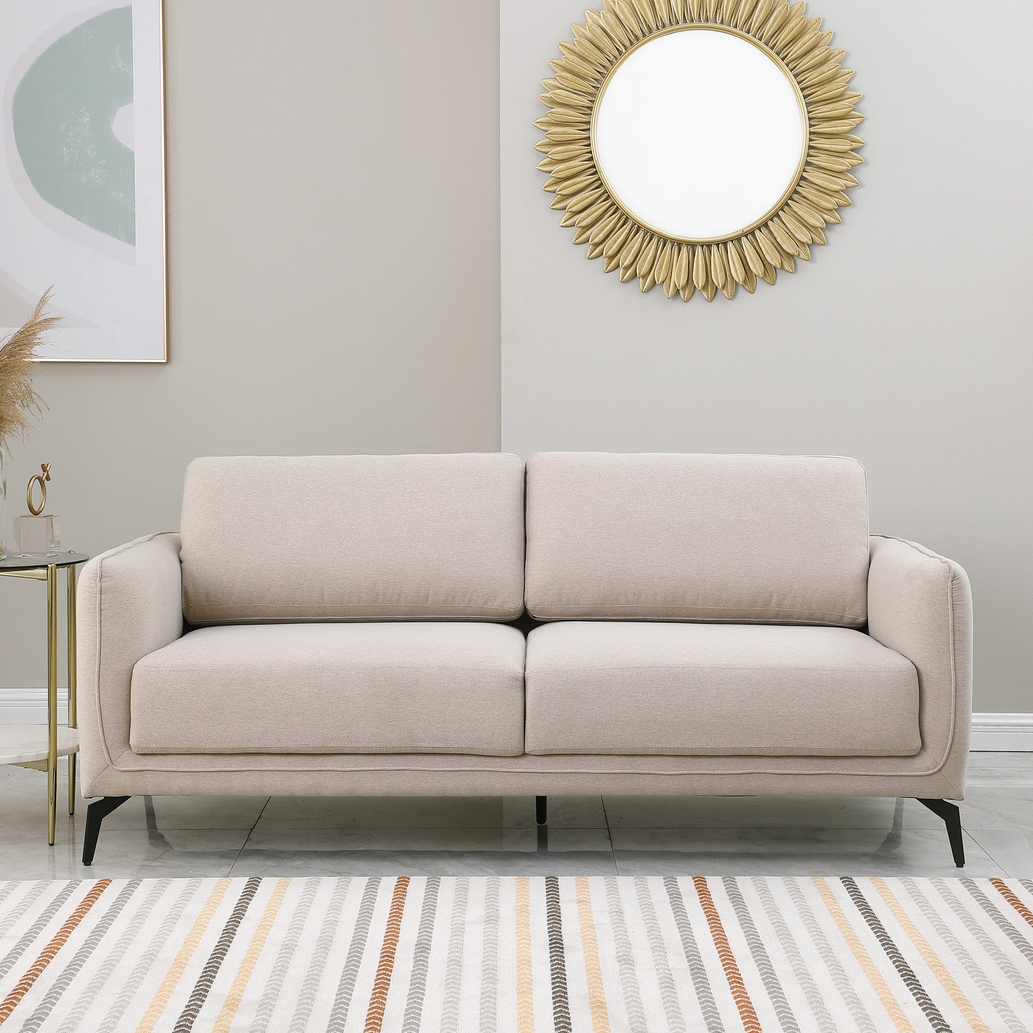 Beige upholstered sofa with black metal legs in a living room, featuring a round sunburst mirror and a side table with flowers.