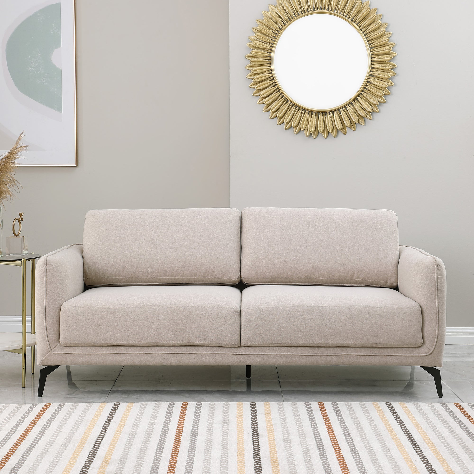 Beige upholstered sofa with black metal legs in a living room, featuring a round sunburst mirror and a side table with flowers.