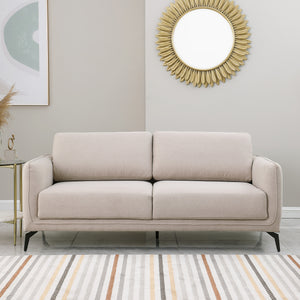 Beige upholstered sofa with black metal legs in a living room, featuring a round sunburst mirror and a side table with flowers.