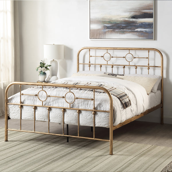 Brass-coloured metal bed frame with circular designs and vertical bars, white bedding with a plaid throw, and pillows. A painting and a lamp with flowers are in the background.