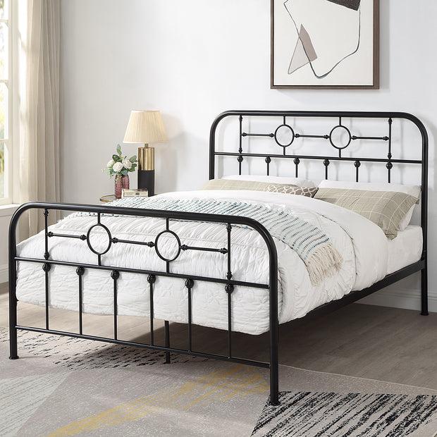 Black metal bed frame with circular designs and vertical bars on the headboard and footboard. The bed has white bedding with a patterned throw and pillows. A modern painting, lamp, and window with curtains are in the background.