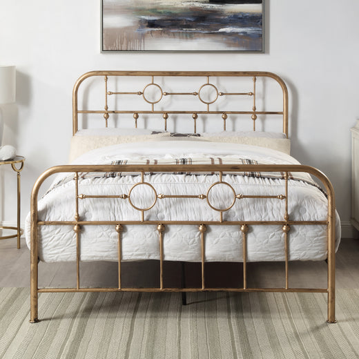 A brass-coloured metal bed is adorned with white bedding, decorative pillows, and a plaid throw blanket. A painting hangs above the bed on a white wall.