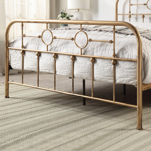 Brass-coloured metal bed frame with circular designs and vertical bars at the foot of a bed with a white quilt, and a beige carpet and white lamp in the background.
