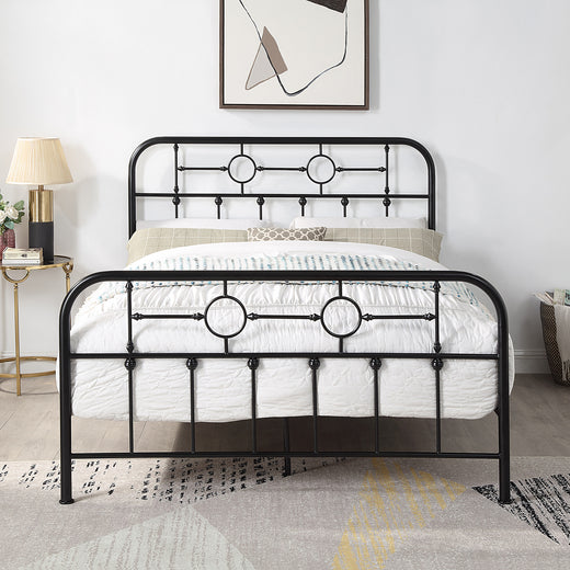 Black metal bed frame with circular designs and vertical bars, white bedding with a patterned throw, and pillows. A modern painting and a lamp with flowers are in the background.