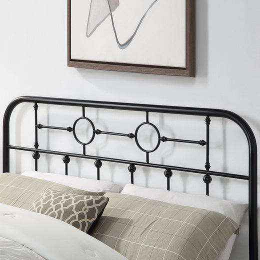 Close-up of a black metal headboard with circular designs and vertical bars. The bed is made with patterned pillows and white bedding. An abstract painting is visible above the headboard.