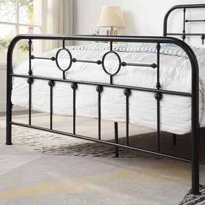 Close-up of a black metal bed frame with circular designs and vertical bars at the foot of the bed. White bedding with a patterned throw is visible. A lamp and window are in the background.