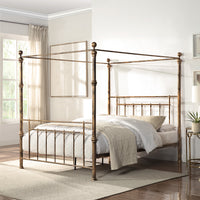 A brass-coloured metal four-poster bed with finials and vertical rods, positioned centrally against a light wall, next to framed artwork and a bedside table.