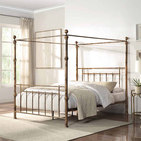 A brass-coloured metal four-poster bed with finials and vertical rods, positioned centrally against a light wall, next to framed artwork and a bedside table.