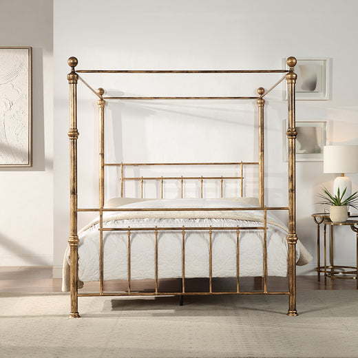 A brass-coloured metal four-poster bed with finials and vertical rods, positioned centrally against a light wall, next to framed artwork and a bedside table.